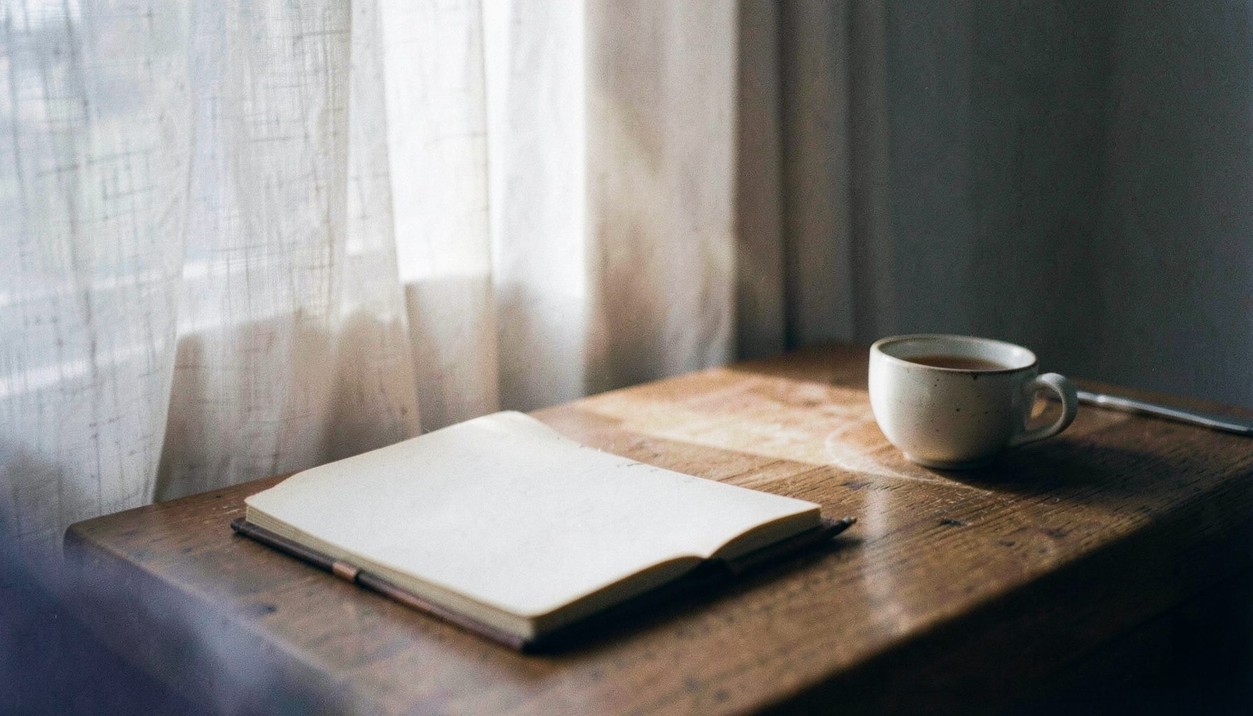 Soft daylight pouring through a pale linen curtain onto a wooden desk, with a single open notebook and a cup of tea, in muted lavender and warm cream tones